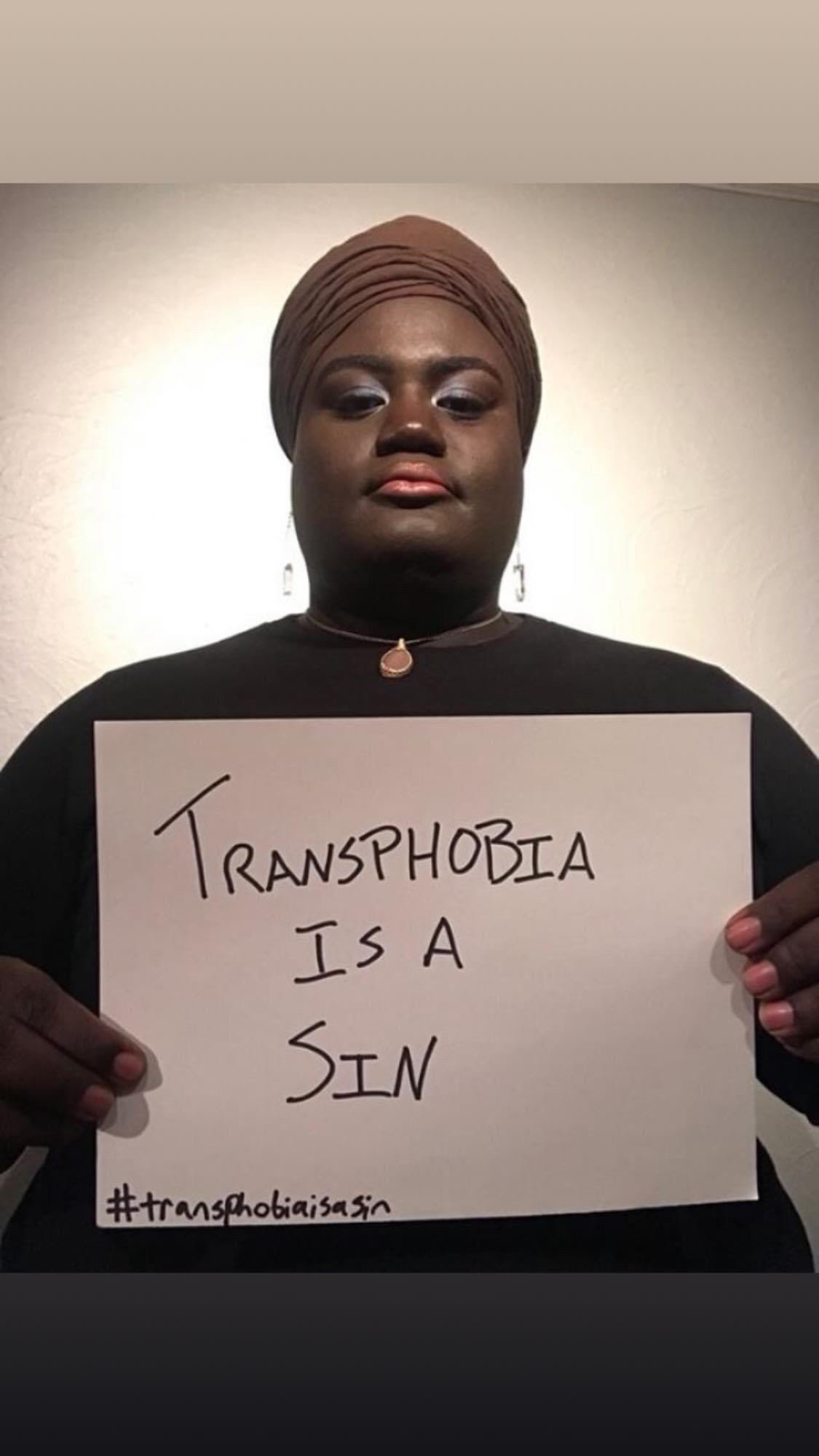 Dark skinned Black Transfemme person with a full beat face staring into the camera while baring a sign declaring Transphobia Is a Sin
