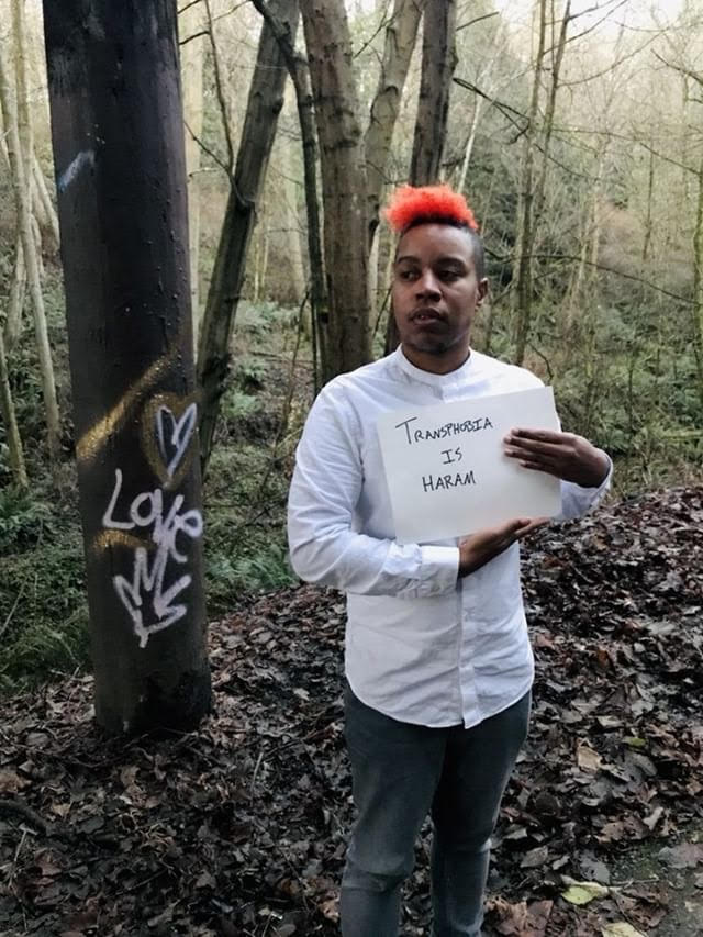 Light skinned Black Transmasc person with fire red hair holding a sign that says Transphobia is Haram in a wooded area