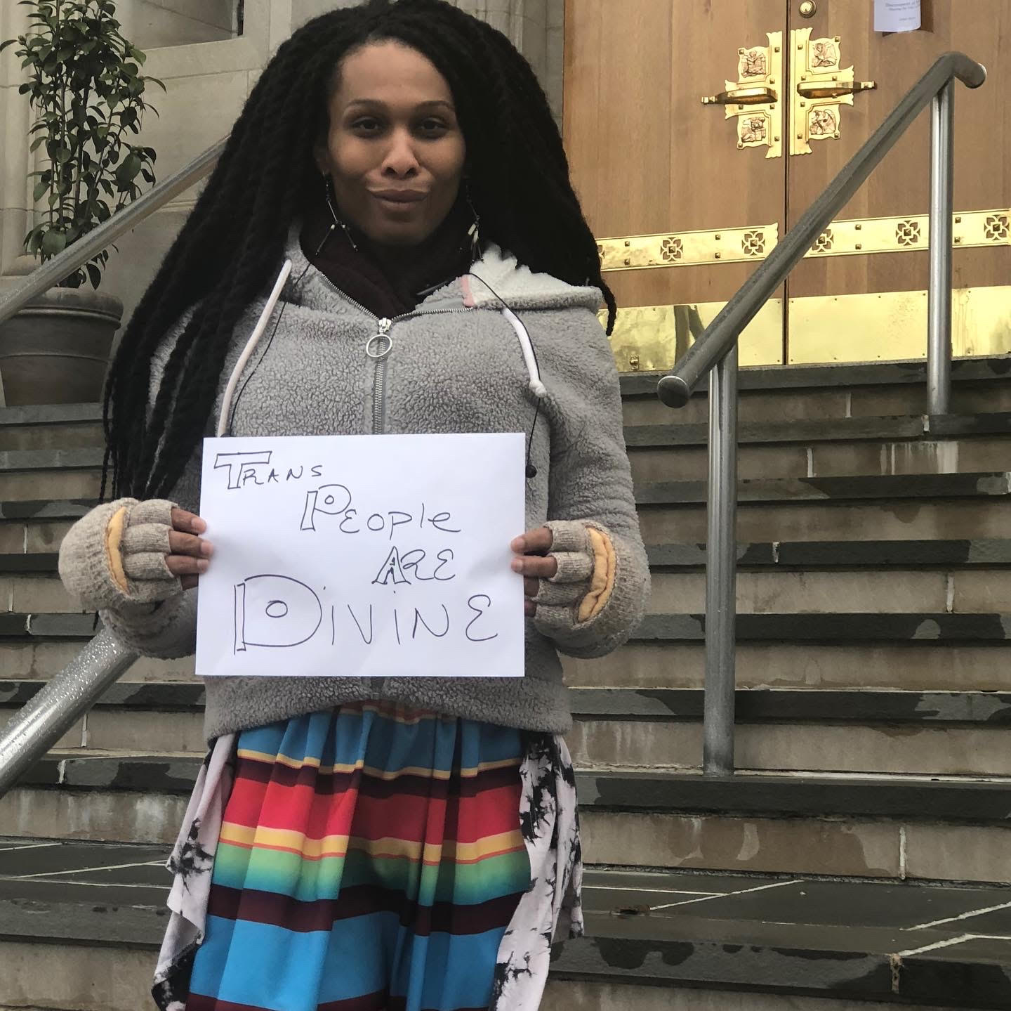 Brown skinned Black Trans Woman in front of a church holding a sign that reads Trans People are Divine and wearing a multicolored striped skirt with grey sweatshirt.